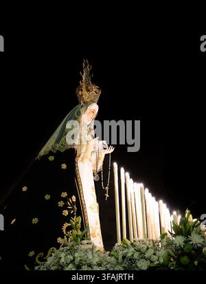 Statue der Jungfrau Maria steht nachts groß. Kerzen leuchten hinter ihr und beleuchten ihre Gewänder. Der Schleier fällt anmutig über ihre Schultern. Blumen umgeben Stockfoto