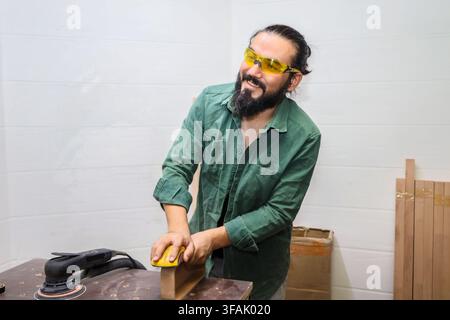 SCHREIBER ARBEITET AN DER HOLZARBEITSBANK, SCHLEIFT MIT DER HAND EIN GEKRÜMMTES HOLZSTÜCK, WÄHREND EIN ELEKTROSCHLEIFER IN DER NÄHE RUHT, TRÄGT SICHERHEITSBRILLEN IN HELLEM WERKSTATT MIT S Stockfoto