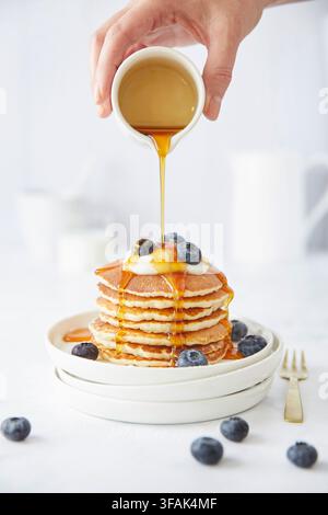 Ein Teller mit Pfannkuchen und Blaubeeren. Von oben gießt eine Hand Ahornsirup über die Pfannkuchen. Eine gemütliche Küchenatmosphäre mit einer fröhlichen Stimmung. Stockfoto