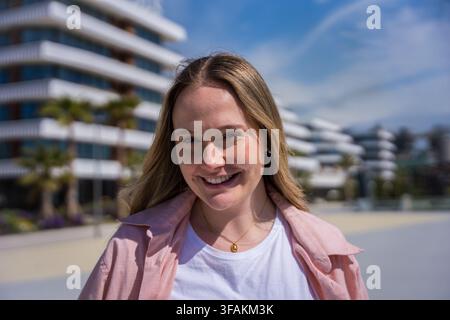 Lächelnde junge blonde Frau posiert im Freien in einer sonnigen modernen Nachbarschaft Stockfoto