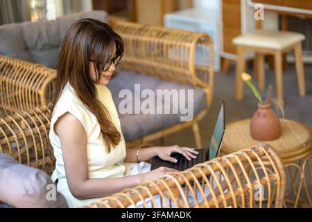 Junge Frau mit Brille, die an einem Laptop in einem komfortablen modernen Interieur arbeitet, repräsentiert Remote-Arbeit, Freelancing, Produktivität, digitale b Stockfoto