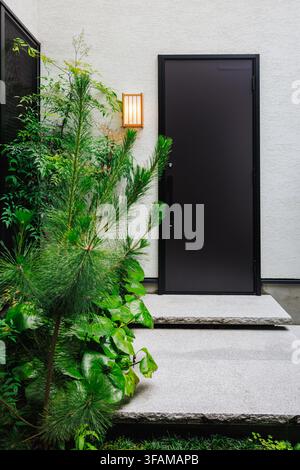 Minimalistischer japanischer moderner Hauseingang mit dunkler Tür, Stein Stufen, leuchtender Laterne und üppiger Kiefernpflanze Stockfoto