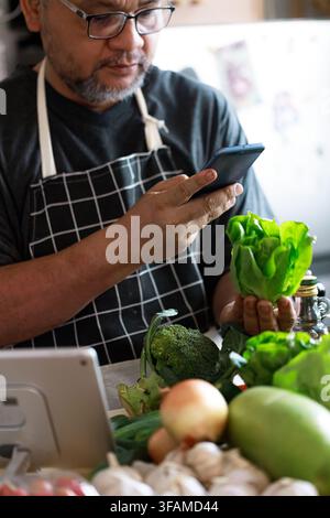 Mittelalterer asiatischer Mann verwendet ein Smartphone, während er zu Hause frisches Gemüse auswählt. Konzept von gesundem Altern, Entscheidungsfindung bei der Ernährung, präventiver Gesundheit Stockfoto