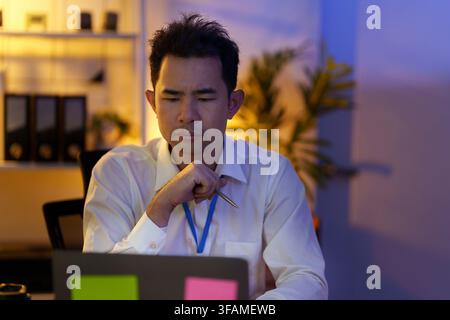 Konzentrierter asiatischer Geschäftsmann denkt, während er spät im Büro am Laptop arbeitet Stockfoto