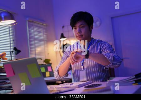 Asiatischer Geschäftsmann überprüft Smartwatch während der Spätschicht im Büro Stockfoto