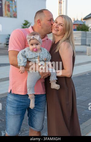 ein herzerwärmender Familienmoment, bei dem ein Vater und eine Mutter ihr Baby liebevoll küssen und halten, in einer urbanen Umgebung im Freien, in einem Nahaufnahme aufgenommen Stockfoto