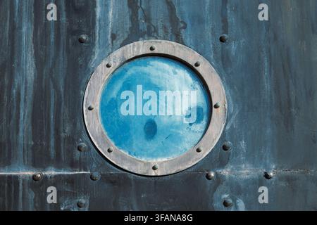 Nahaufnahme eines antiken Stahlgefäßüberzugs mit Nieten, Rostflecken und einem blauen Glasbullauge, Konzept von Meeresabenteuer und Unterwassererkundung Stockfoto
