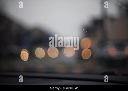 Abstrakte Hintergrundansicht durch eine nasse Windschutzscheibe eines Autos mit Regentropfen und weichen, verschwommenen Bokeh-Lichtern vom Stadtverkehr während eines regnerischen Abends in gl Stockfoto