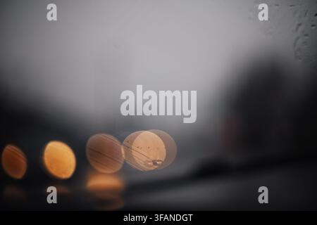 Abstrakte Nahaufnahme durch eine regnerische Autoscheibe, die verschwommene warme Lichter und weiche Bokeh-Formen bei trübem und nassem Wetter zeigt Stockfoto