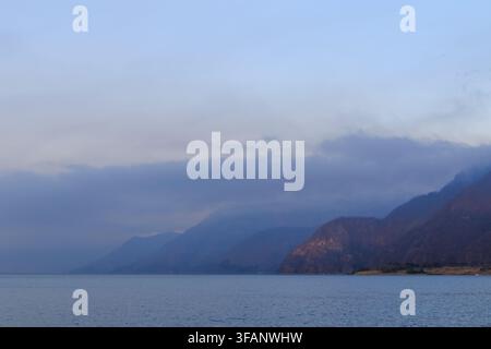 Ruhiges Seewasser erstreckt sich zu dunklen Berghängen unter geschichteten blaugrauen Wolken in einer stillen nebligen Abendlandschaft. Stockfoto