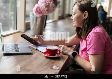 Frau benutzt Smartphone und Laptop in einem Café mit Kaffee Stockfoto