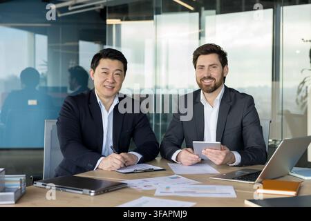 Verschiedene Geschäftspartner lächeln, schauen in die Kamera und arbeiten gemeinsam an einem Schreibtisch in einem modernen Firmenbüro zusammen, was erfolgreiche Teamarbeit und professionelle Partnerschaft zeigt Stockfoto