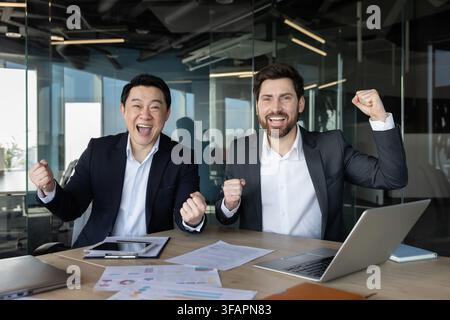 Zwei verschiedene Geschäftsleute in Anzügen feiern einen großen Erfolg an einem modernen Bürotisch, jubeln und stemmen die Fäuste in die Luft über Laptops und Dokumenten, um Teamwork und Erfolg hervorzuheben Stockfoto