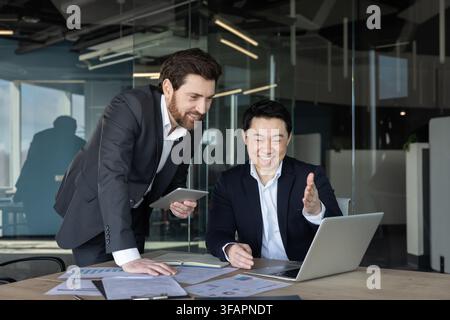 Zwei verschiedene Geschäftsleute arbeiten in einem modernen Büro zusammen, ein Mann steht mit einem Tablet und der andere Mann sitzt an einem Schreibtisch mit einem Laptop, beide diskutieren über die Arbeit und zeigen Teamarbeit Stockfoto