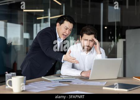 Wütender asiatischer Chef schreit und gestikuliert intensiv gegenüber einem gestressten kaukasischen Mitarbeiter, der niedergeschlagen an einem Laptop in einem modernen Büro sitzt, und stellt Arbeitsplatzkonflikte und Druck dar Stockfoto
