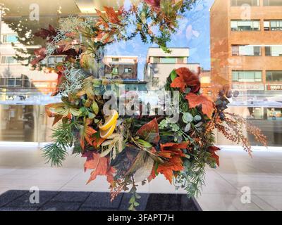 Ein aus verschiedenen Blättern und Blumen gefertigter Kranz ist in einem Schaufenster sichtbar, mit einer städtischen Kulisse, die Gebäude und Geschäfte umfasst. Stockfoto