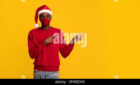 African Guy In Schützende Gesichtsmaske Und Weihnachtsmann Hut Zeigen Zur Seite Stockfoto