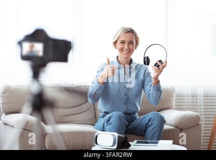 Junge Influencerin, die zu Hause Videoinhalte aufzeichnet, mit modernen Kopfhörern review macht und den Daumen nach oben zeigt Stockfoto
