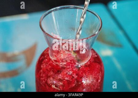 eine gläserne Karaffe gefüllt mit einem lebendigen, erfrischenden zerkleinerten roten Beeren-Eisgetränk und einem verdrehten Metallbarschlöffel, der auf einem blauen Café-Tisch ruht. Stockfoto