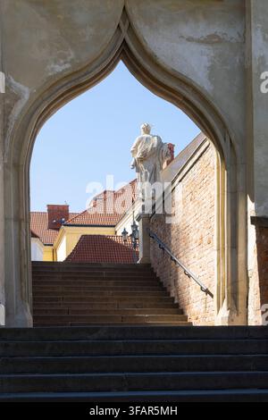 Brno, Tschechische Republik. 12. August 2026. Eine steinerne Treppe führt zu einem gewölbten Tor. Durch das Gewölbe steht eine Statue einer Gestalt in Gewändern auf einem r Stockfoto