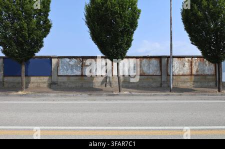Bäume auf dem Bürgersteig und Betonmauer mit leeren Metalltafeln für Straßenwerbetafeln dahinter. Himmel darüber, große Straße davor. Hintergrund Stockfoto