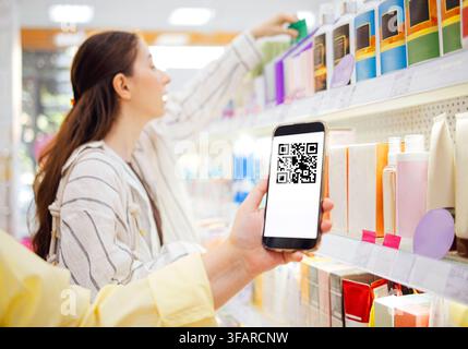 Frau betrachtet Kosmetikprodukte auf Regalen, während sie ein Smartphone mit einem QR-Code in einer Einzelhandelsumgebung hält Stockfoto