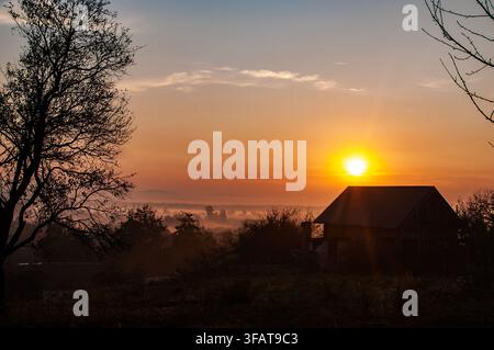 Die helle aufgehende Sonne scheint über ein kleines Landhaus, das sich gegen ein nebliges Tal im Morgengrauen abhebt. Stockfoto