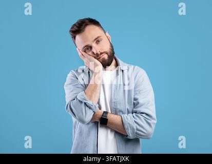 Gelangweilter junger Kerl, der sich auf seine Hand lehnte und ansah Kamera über blauem Studiohintergrund Stockfoto