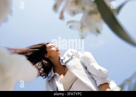 Junge Frau steht mit nach hinten geneigtem Kopf gegen einen blauen Himmel, umgeben von blühenden weißen Blumen, aus niedriger Perspektive aufgenommen Stockfoto