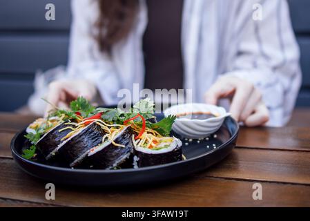Sushi-Rollen, die auf einem schwarzen Teller mit einer kleinen Schüssel Dippingsauce serviert werden, garniert mit Kräutern und Gemüse, gehalten von einer Person in einem lässigen Esszimmer Stockfoto