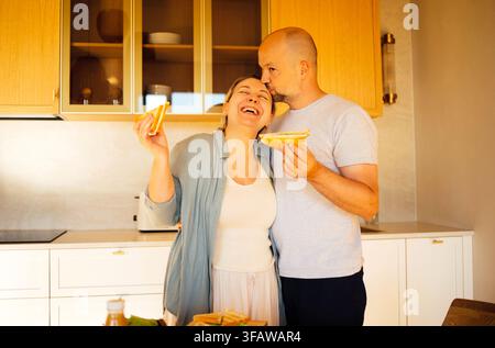 Paar in einer modernen Küche teilt einen leichten Moment, während es Sandwiches hält, mit Holzkabinetten und einer Arbeitsplatte im Hintergrund Stockfoto