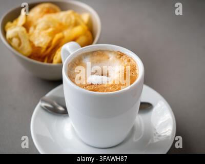 Heißer Kaffee mit cremiger Schaumhaube und Zuckerkörnern, serviert mit einer Schüssel Kartoffelchips Stockfoto