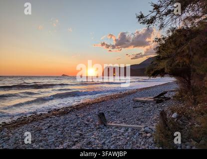 Die untergehende Sonne neigt sich zum Horizont und wirft einen glühenden orangefarbenen Schimmer über sanfte Meereswellen, die an einem felsigen Kieselstrand brechen. Dunkelgrüne Bäume und w Stockfoto