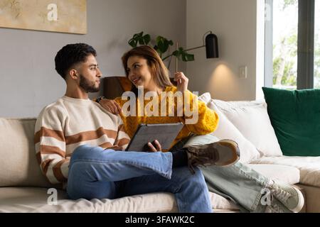 Afroamerikanisches Paar sitzt auf beigem Sofa im Wohnzimmer, trägt Pullover und hält ein Tablet Stockfoto