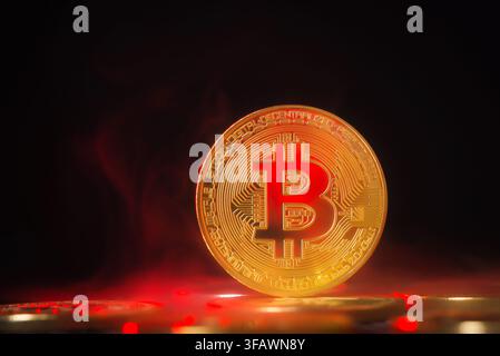 Bitcoin-Kryptowährungsmünze, die aufrecht auf einer reflektierenden Oberfläche steht, beleuchtet von dramatischem rotem Neonlicht und Rauch. Stockfoto