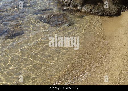 Eine Hochwinkelaufnahme zeigt klares, türkisfarbenes Wasser mit kaustischen Lichtmustern, die über nassen Sand und dunkle Küstenfelsen in hellem Sonnenlicht fließen. Stockfoto