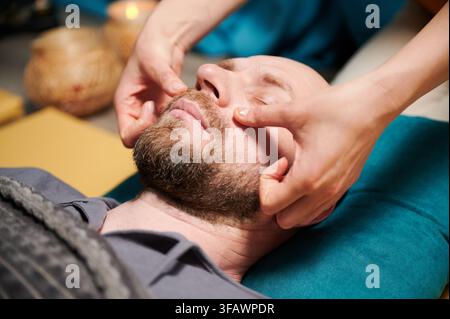 Nahaufnahme eines Mannes, der eine thai-Gesichtsmassage bekommt. Masseurin, die traditionelle Massagebehandlung für männlichen Patienten durchführt. Glatzköpfige Kundin mit geschlossenen Augen, Meditation, Handmassage in ruhiger Atmosphäre. Stockfoto
