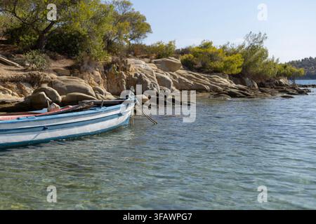 Ein traditionelles blau-weißes Holzboot ruht an einem felsigen Ufer. Das klare türkisfarbene Wasser trifft auf eine sonnenbeschienene Küste mit Bäumen und großen Felsbrocken. Stockfoto