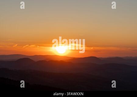 Leuchtende Sonne, die hinter einem fernen Gebirgskamm unter einem warmen orangefarbenen Himmel untergeht, mit dunklen, geschichteten Hügeln im Vordergrund. Konzept des Tagesendes, der Ruhe, der Natur Stockfoto