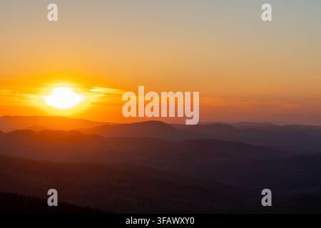 Strahlender orangefarbener Sonnenuntergang über geschichteten Bergsilhouetten mit leuchtender Sonne über fernen Kämmen. Konzept der dramatischen natürlichen Schönheit, friedlicher Abend Stockfoto