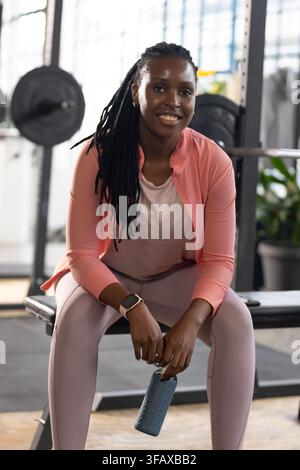 Afrikanische Frau sitzt auf einer Bank im Fitnessstudio in Rosa und hält eine Flasche neben einer mit Gewichten beladenen Hantelstange mit einer Smartwatch Stockfoto