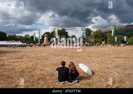 Brecon, Wales, 21. August 2026  Ein junges Paar sitzt zusammen auf einer Decke in einem großen, trockenen Grasfeld und blickt über das überfüllte Gelände des Green Man Festivals unter einem dramatischen bewölkten Himmel. Tag Zwei des Green Man Festivals 2026 in Brecon, Wales. Foto: Rob Watkins/Alamy Live News. INFO: Nach Monaten von Hitze und Dürre gab es am zweiten Tag des Green Man Festivals 2026 eine Mischung aus Sonnenschein und heftigen Regenschauern. Die Menge genoss die erfrischend kühle Wetteränderung. Stockfoto