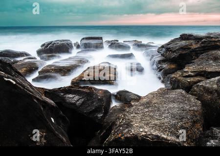 Meereslandschaft aus geschichteten Felsen, umgeben von weichem, nebligem weißem Wasser mit einem türkisfarbenen Ozean und einem blassrosa Horizont. Manly, Australien. Stockfoto