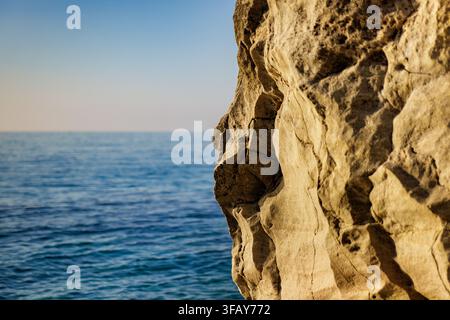 Eine strukturierte, sonnenbeschienene Felswand, die über einen ruhigen blauen Ozeanhorizont blickt, Tropea, Italien. Stockfoto