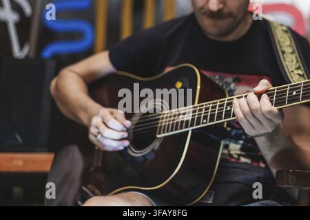 Ein junger kaukasischer Mann mit Bart spielt eine Akustikgitarre. Er trägt ein schwarzes T-Shirt und konzentriert sich in einer lockeren Umgebung auf seine Musik Stockfoto