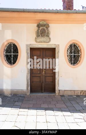 Brno, Tschechische Republik. 12. August 2026. Eine zentrale hölzerne Doppeltür ist von zwei ovalen Fenstern an einer hellen Wand flankiert. Über der Tür ein kunstvolles Stockfoto