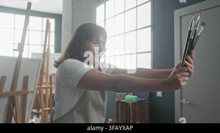 Frau lächelt, hält mit beiden Händen Pinsel, Arme zum Fenster im Kunststudio ausgestreckt, gegenüber Staffeleien und natürlichem Licht; kreative Gelassenheit. Stockfoto