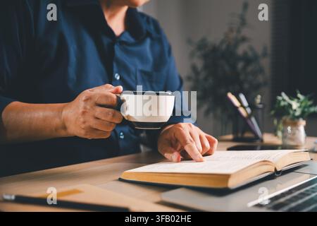 Professioneller hält eine Kaffeetasse und liest ein offenes Buch auf einem Laptop, repräsentiert Selbststudium, Fernlernen, Kaffeepause, lebenslanges Lernen, Stockfoto