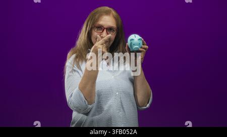 Seniorin mit roter Brille hält einen blauen Sparschwein in der rechten Hand und zeigt mit dem Zeigefinger auf den Münzschlitz im Studio; Rentensparbetrag Stockfoto