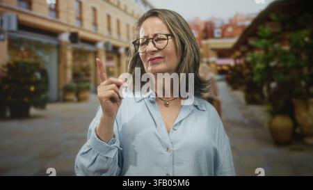 Hispanische Seniorin mit grauem Haar und Brille zeigt mit dem Zeigefinger und blinzelt, während sie in einer Straße steht, die von Geschäften und Pflanzern gesäumt ist; Missbilligung Stockfoto